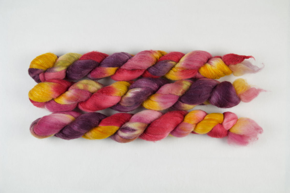Braided fiber skein with purple, red, pink, mustard, and gold on a white background