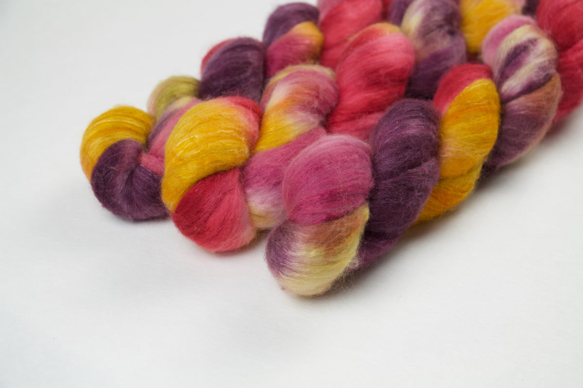Braided fiber skein with purple, red, pink, mustard, and gold on a white background