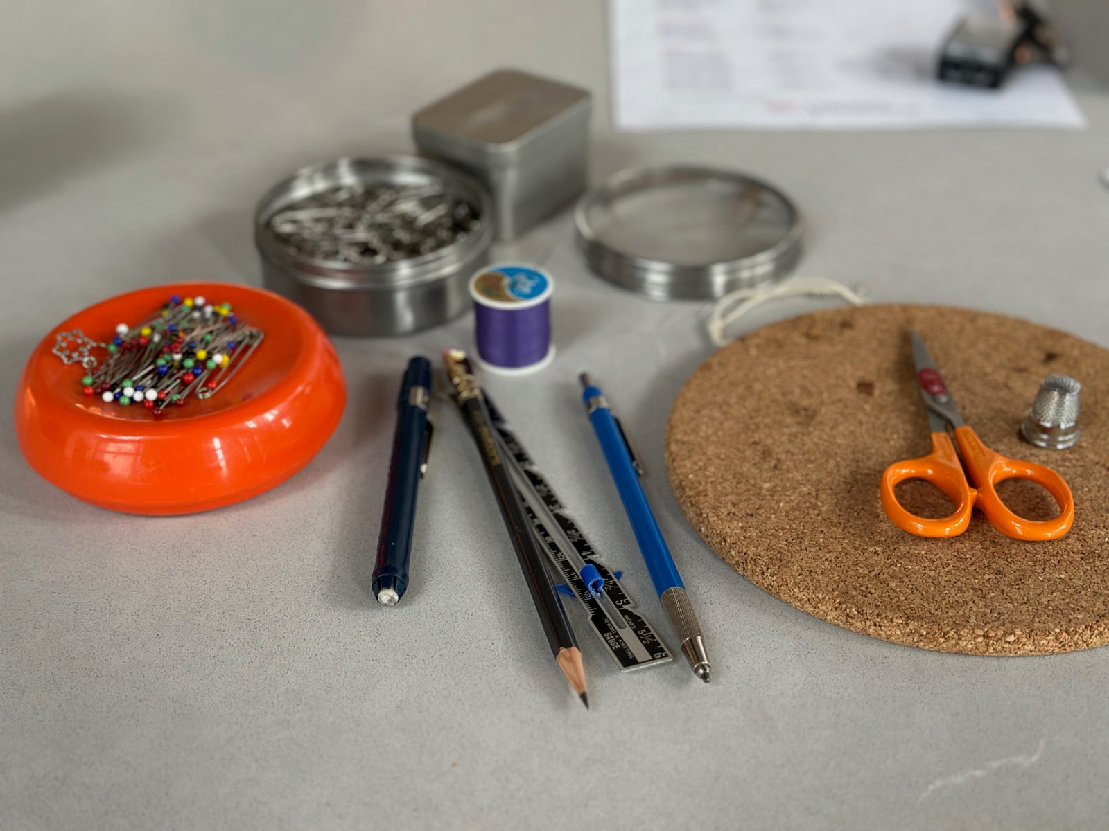 Stationery items including pens, pencils, scissors, and a container of pins on a gray surface.